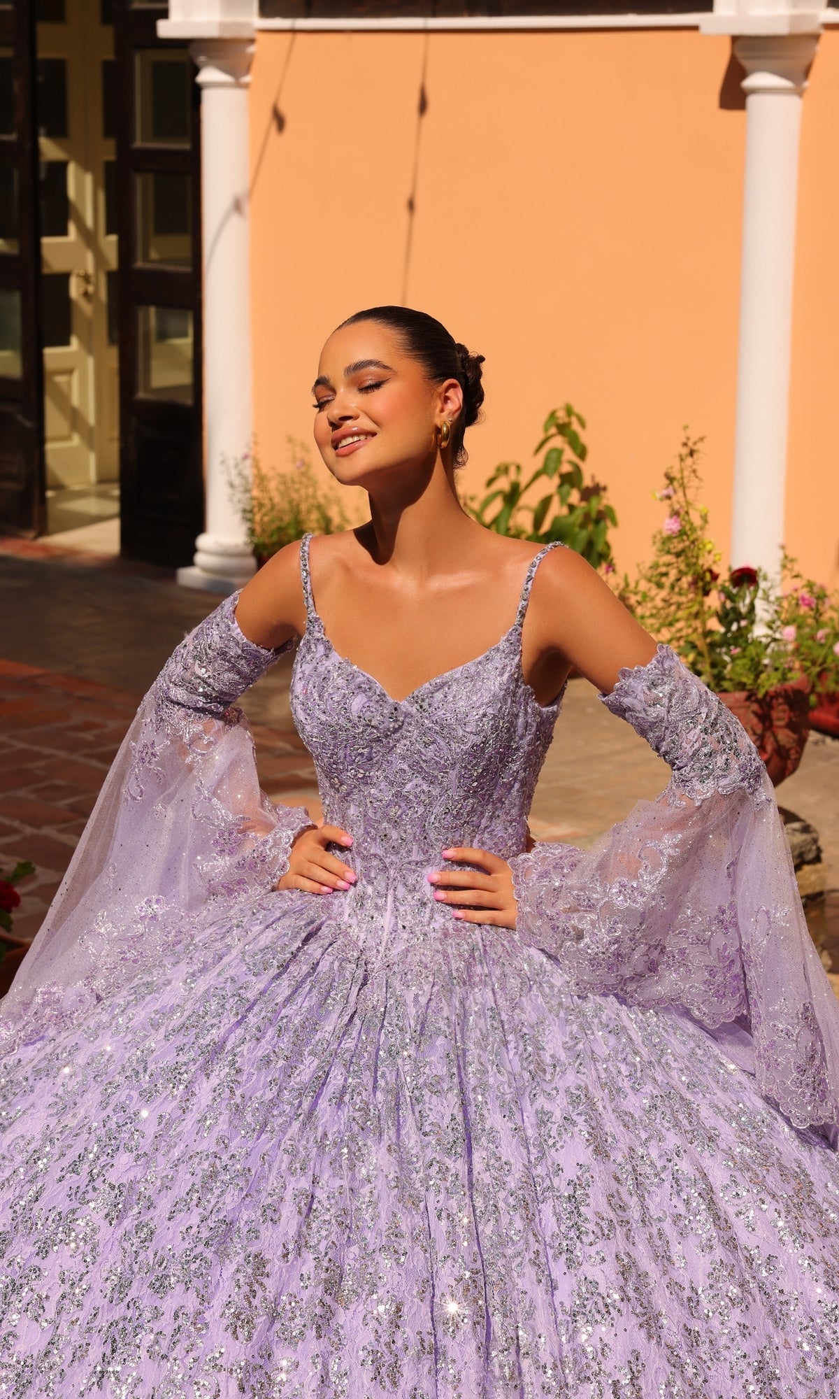 A woman poses outdoors in the sunlight wearing the Quinceanera Dress 54303 By Amarra, a sparkling lavender sequin ball gown with off-the-shoulder sleeves, standing before a peach building with white columns.